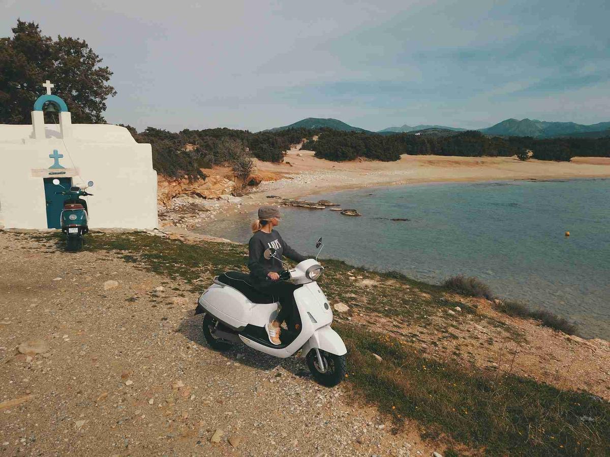 Person Sitting on Scooter Near Beach with Chapel and Sea in Background