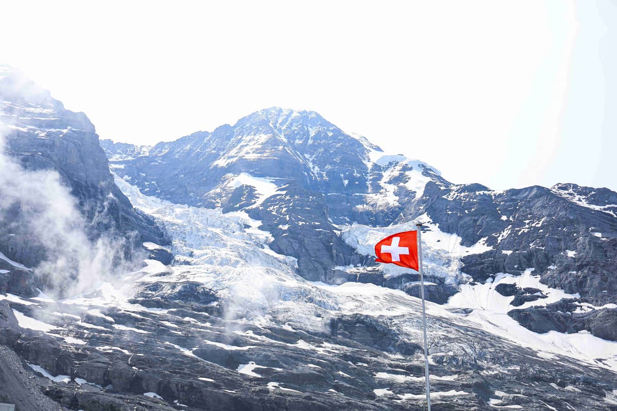 Swiss flag waving before a glacier-covered Alpine peak.