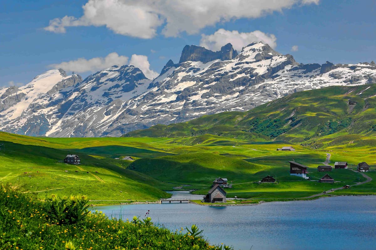 Paisaje alpino con un lago, campos verdes y montañas nevadas.